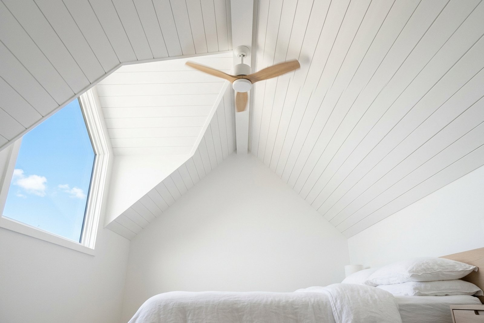 A bright attic bedroom with a sloped white ceiling and a ceiling fan running above a low bed, midday summer light