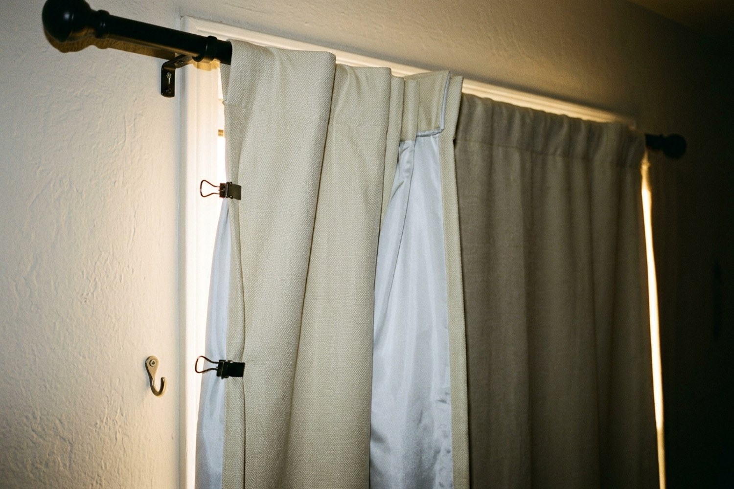 Pale thermal curtain panels overlapping at the middle of a west-facing window, side edges clipped to the wall to seal the side gaps where low afternoon sun would leak in