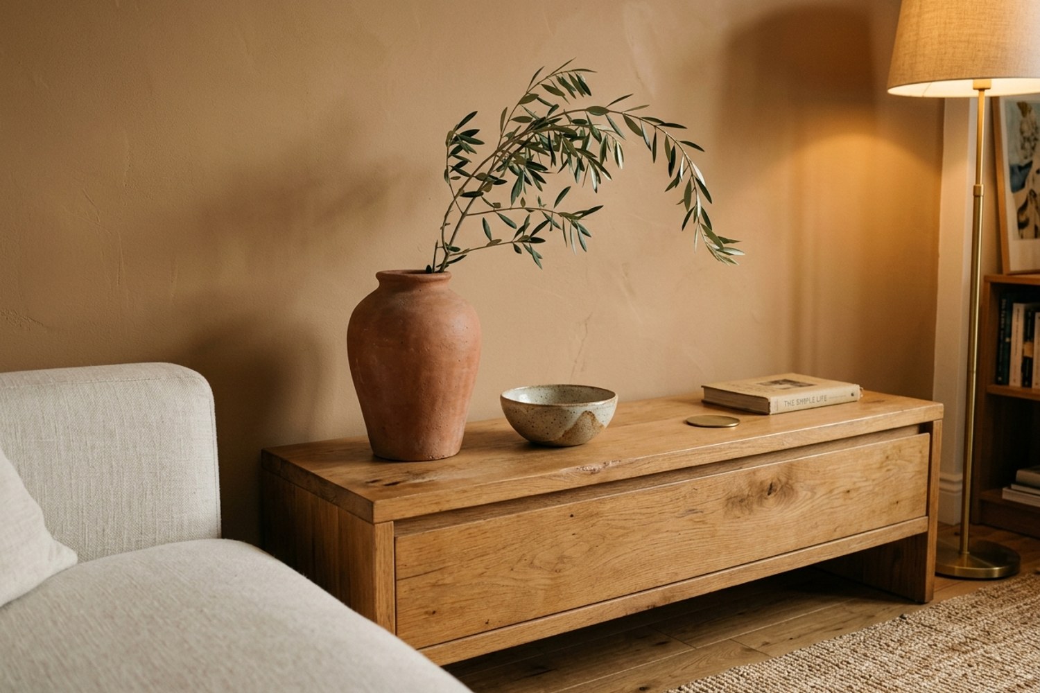 A close-up detail of the 2026 formula: a low oak credenza against a caramel earth-tone wall, one tall terracotta vase with an olive branch, a small ceramic bowl, and warm 2700K lamp light grazing the surface